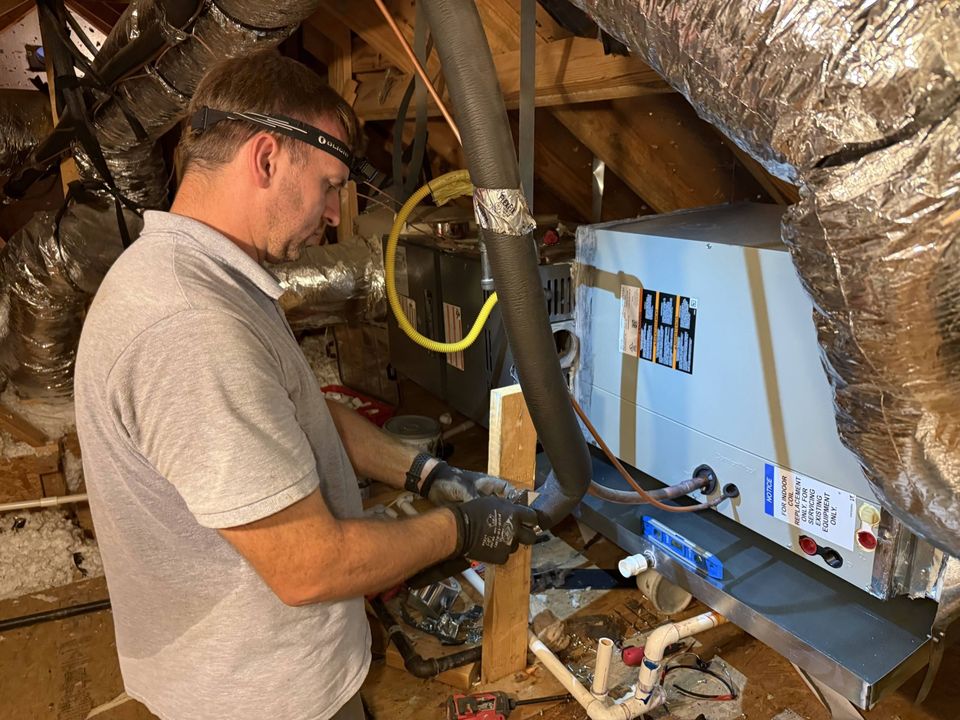 Technician connecting the refrigerant line set at the evaporator coil of an attic furnace, with insulated flex duct runs overhead