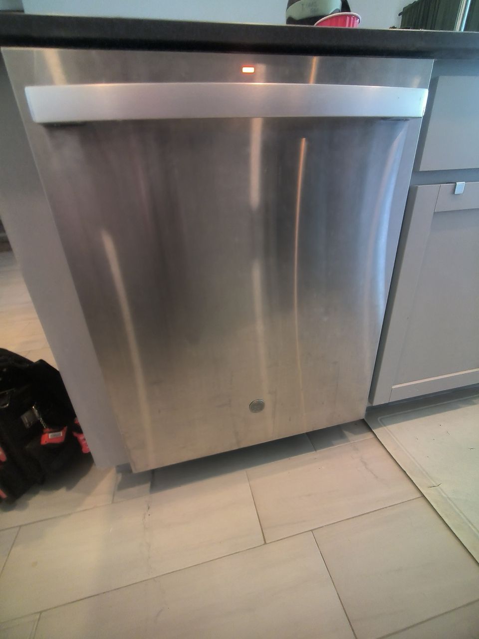 Stainless built-in dishwasher with top-edge controls under a dark countertop, a technician's tool bag on the floor beside it