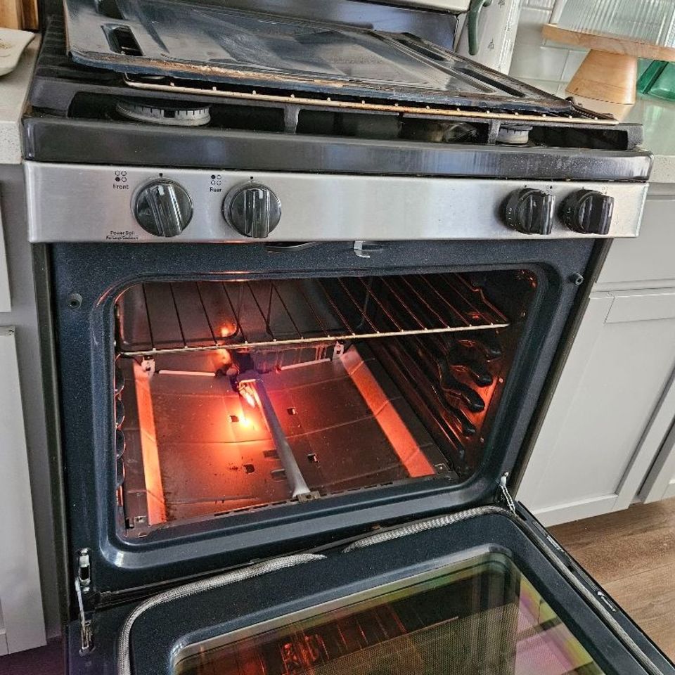 Gas range with the oven door open, the floor panel removed and the bake burner igniter glowing orange